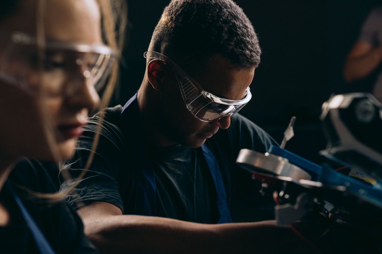 Estudantes usam óculos de proteção e trabalham juntos em um projeto de robótica em uma oficina.