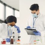 Dois cientistas trabalham em um laboratório, analisando uma amostra ao microscópio e consultando anotações.