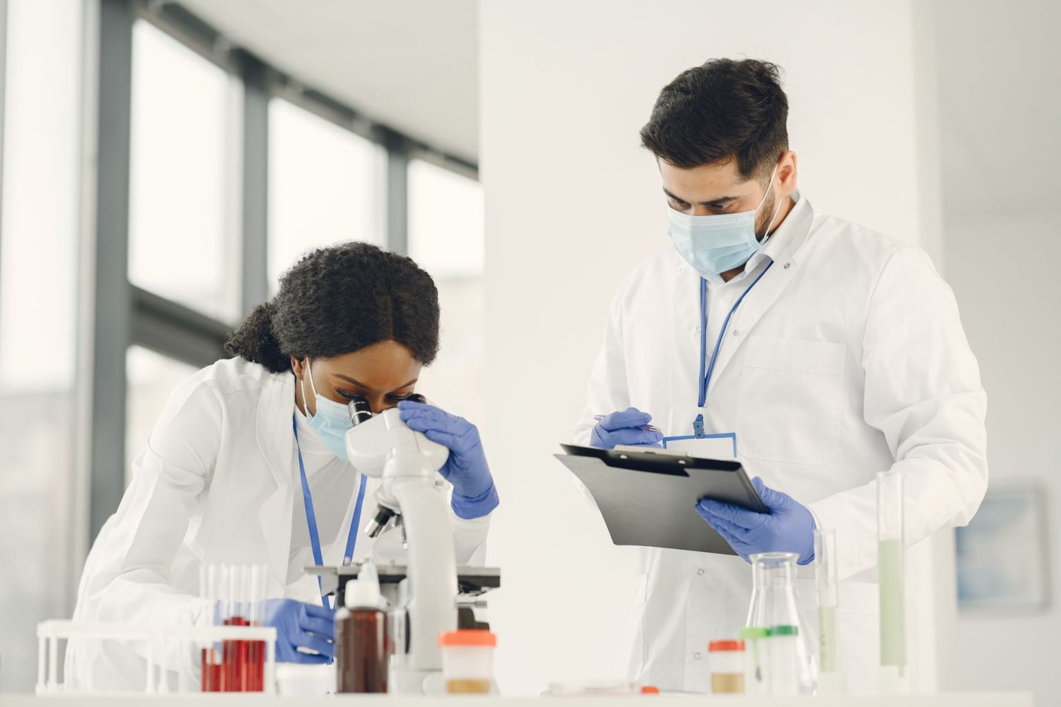 Dois cientistas trabalham em um laboratório, analisando uma amostra ao microscópio e consultando anotações.