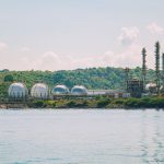 Instalação industrial de armazenamento de gás, com tanques esféricos e chaminés junto ao litoral.