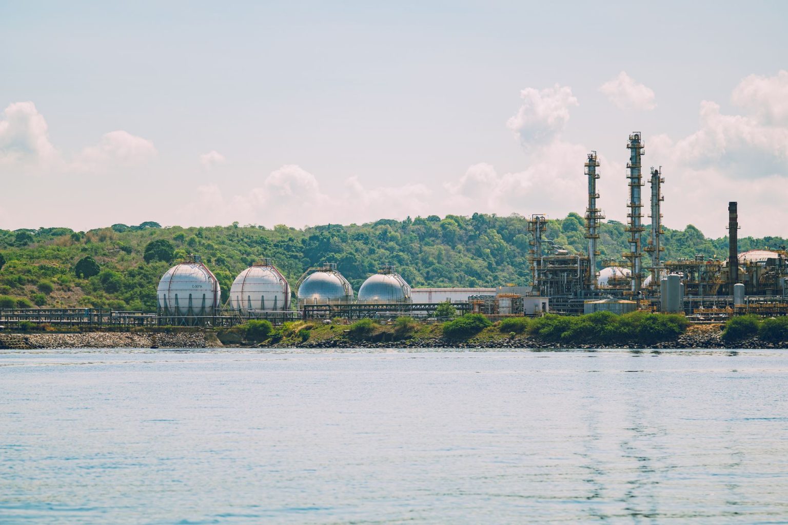Instalação industrial de armazenamento de gás, com tanques esféricos e chaminés junto ao litoral.